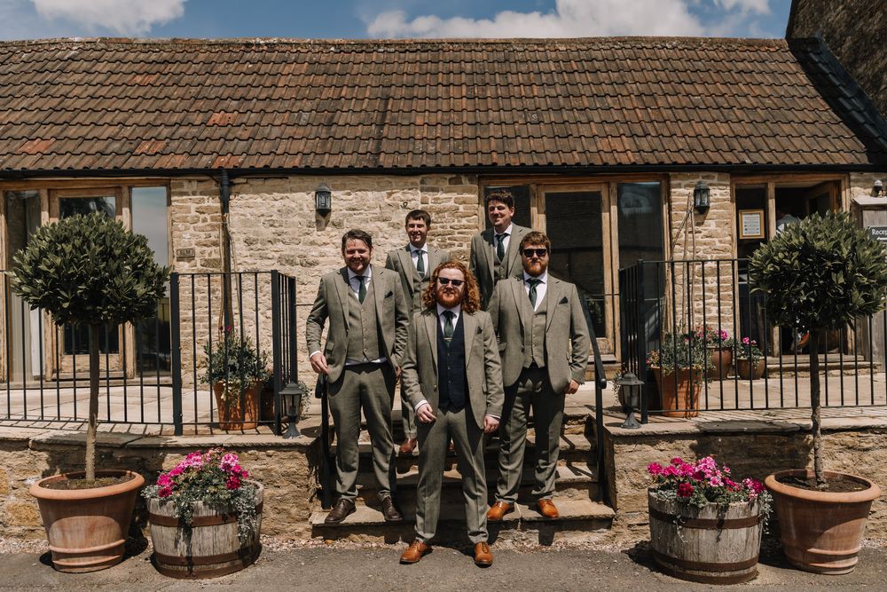 Six groomsmen in matching grey suits pose outside a rustic stone barn venue with potted topiary trees.