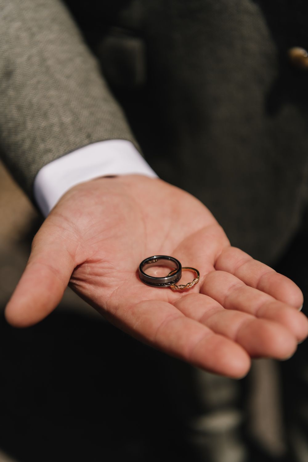 A person in a gray suit holds two wedding rings, one dark and one gold, in the palm of their hand.