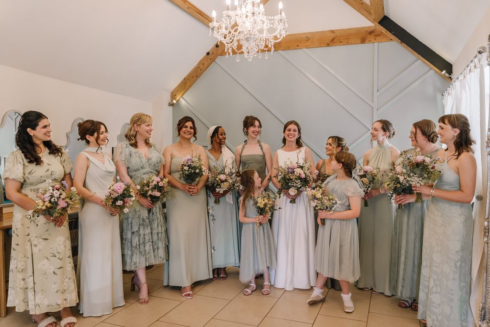 Bridesmaids in sage green and floral dresses holding wildflower bouquets laughing together in a rustic barn wedding venue.