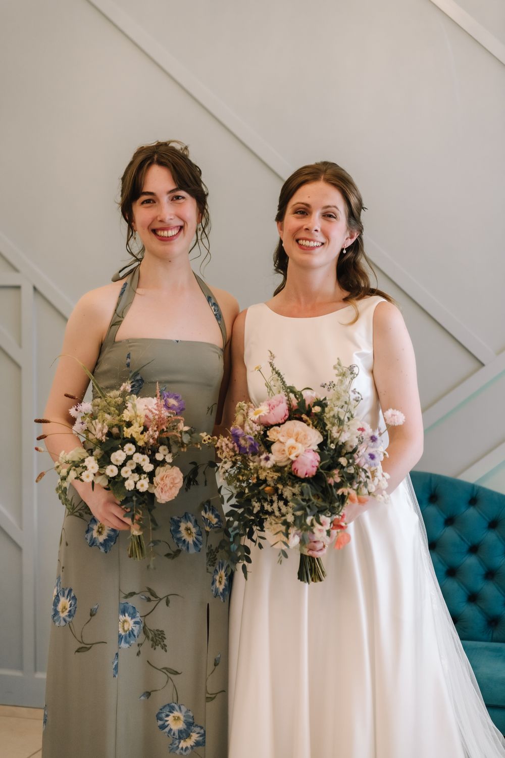 Bride and bridesmaid smiling, holding pastel floral bouquets, wearing white gown and sage floral dress.