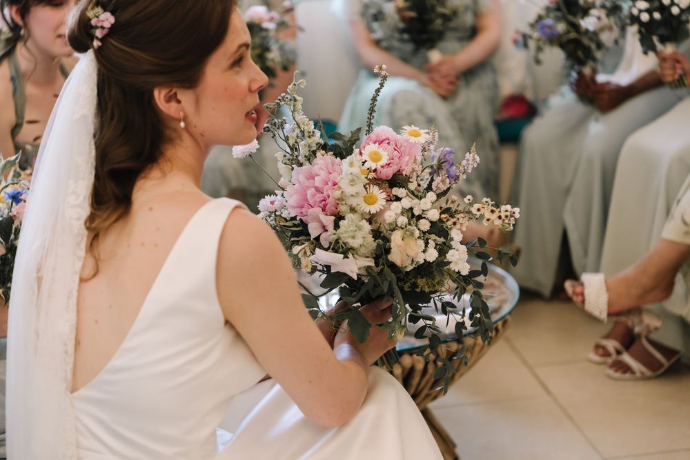 Bride in white gown holding pink and white floral bouquet at wedding ceremony with bridesmaids in background.