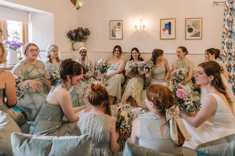 Bridesmaids in sage green dresses sit in a circle holding floral bouquets during wedding preparations.