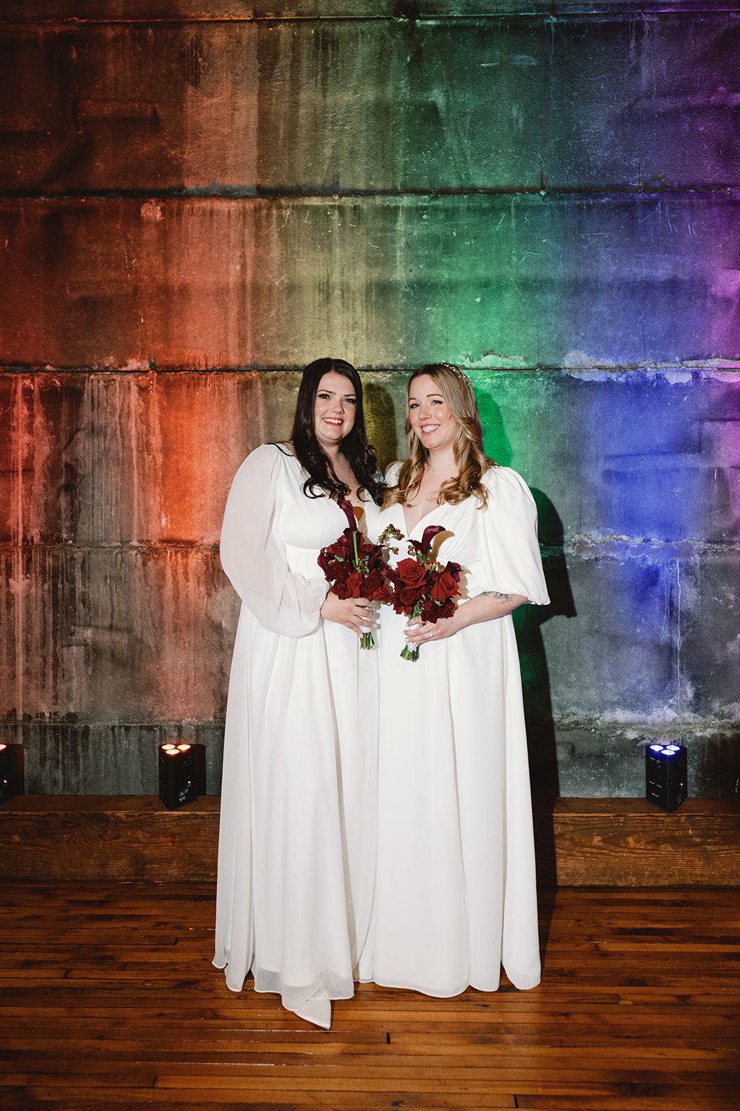 Women in elegant formal dresses pose together at an indoor event with wooden floors and wall decor.