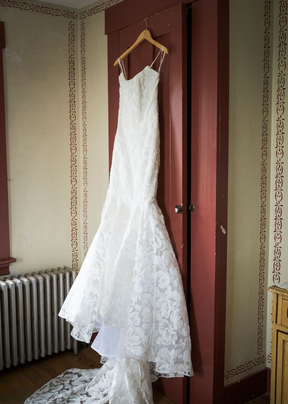 Elegant white lace mermaid wedding dress hanging on a wooden hanger against a red door.