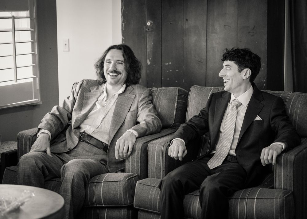 Two men in suits laughing together on armchairs in a black and white photo.