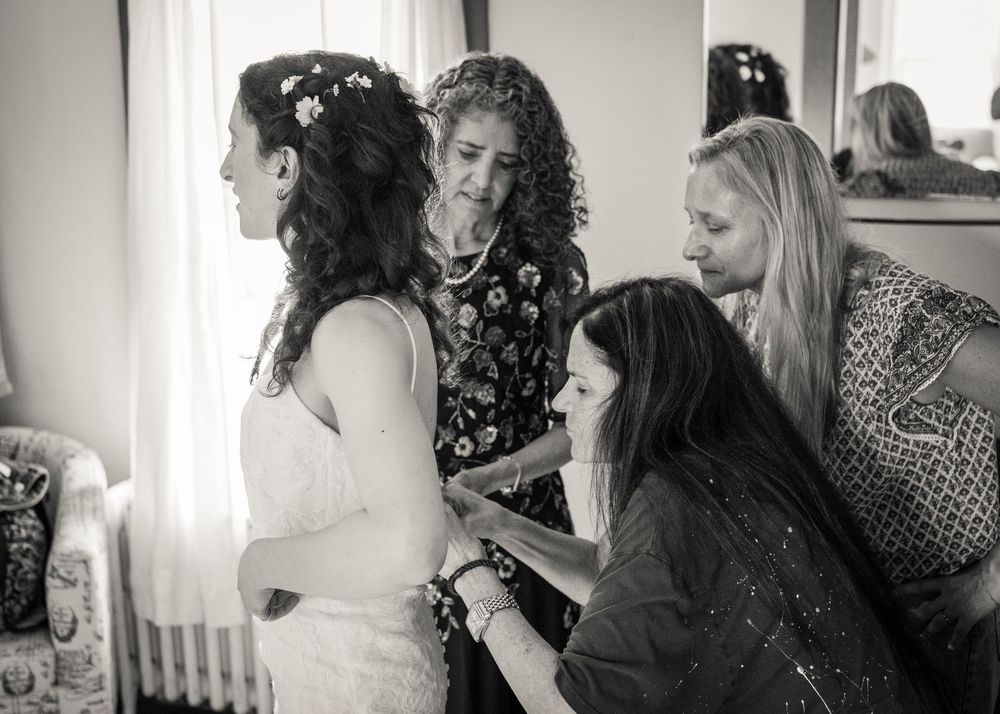 Black and white photo of a bride being helped into her wedding dress by three women in a bright room.