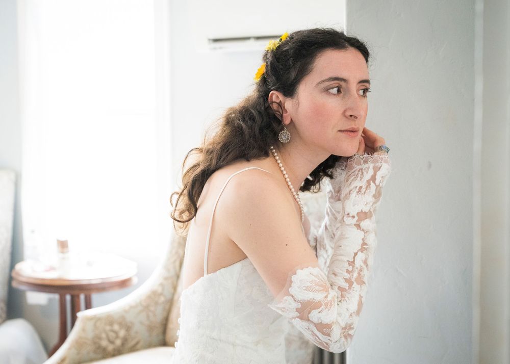 Bride in white lace gown with pearl necklace and yellow hair flower gazes thoughtfully in bright room.