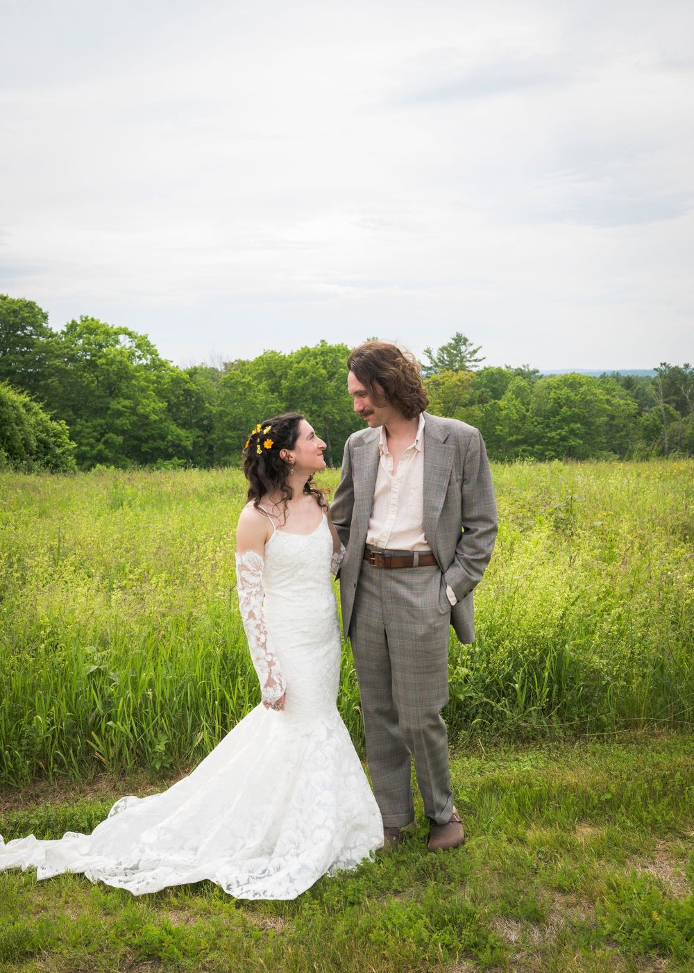 Bride and groom share romantic moment in green meadow, bride in lace mermaid gown with floral hair.