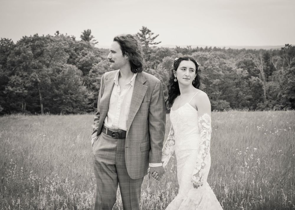 Black and white photo of bride and groom holding hands walking through scenic meadow on wedding day.