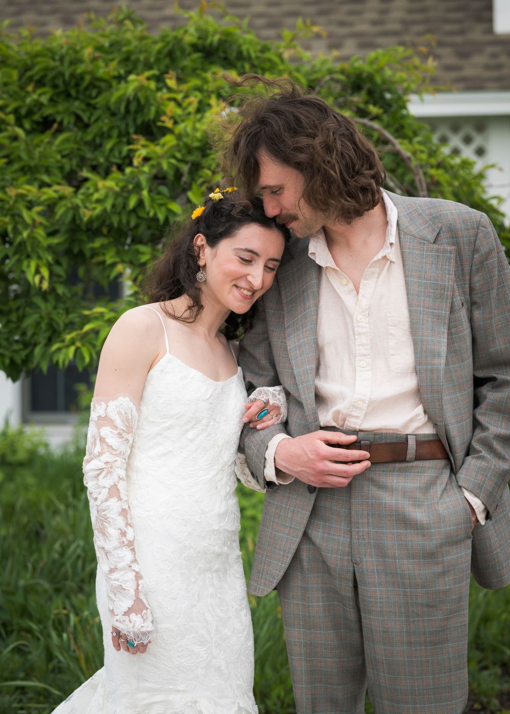 Bride in lace wedding gown and groom in gray suit share a tender moment outdoors.