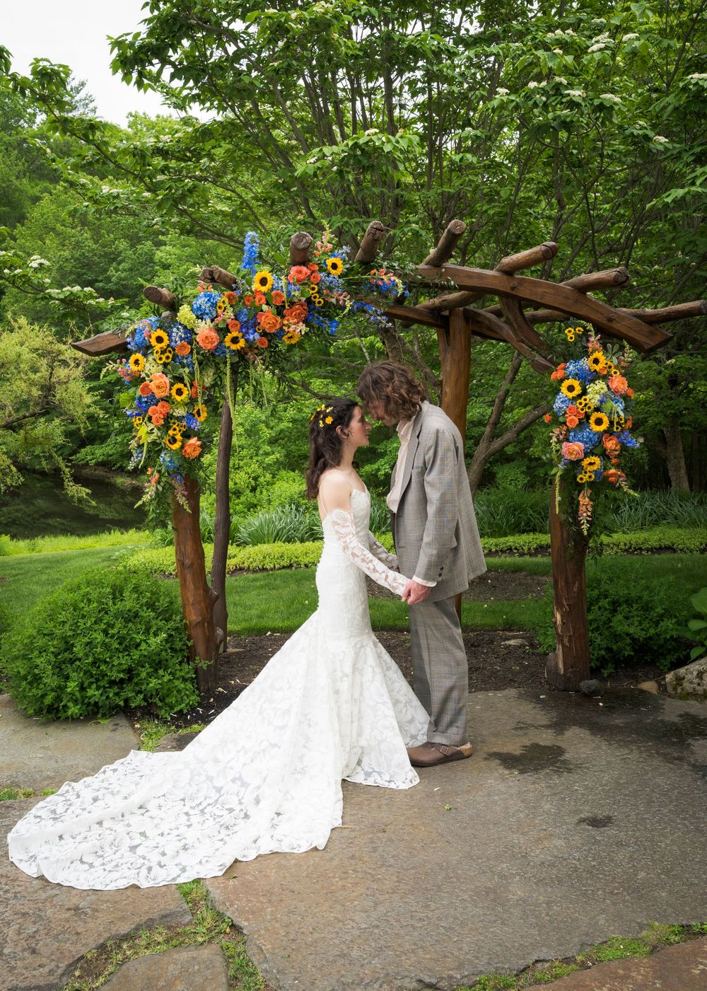 Bride and groom hold hands under a rustic wooden arch adorned with colorful sunflowers and blue flowers in a lush garden.