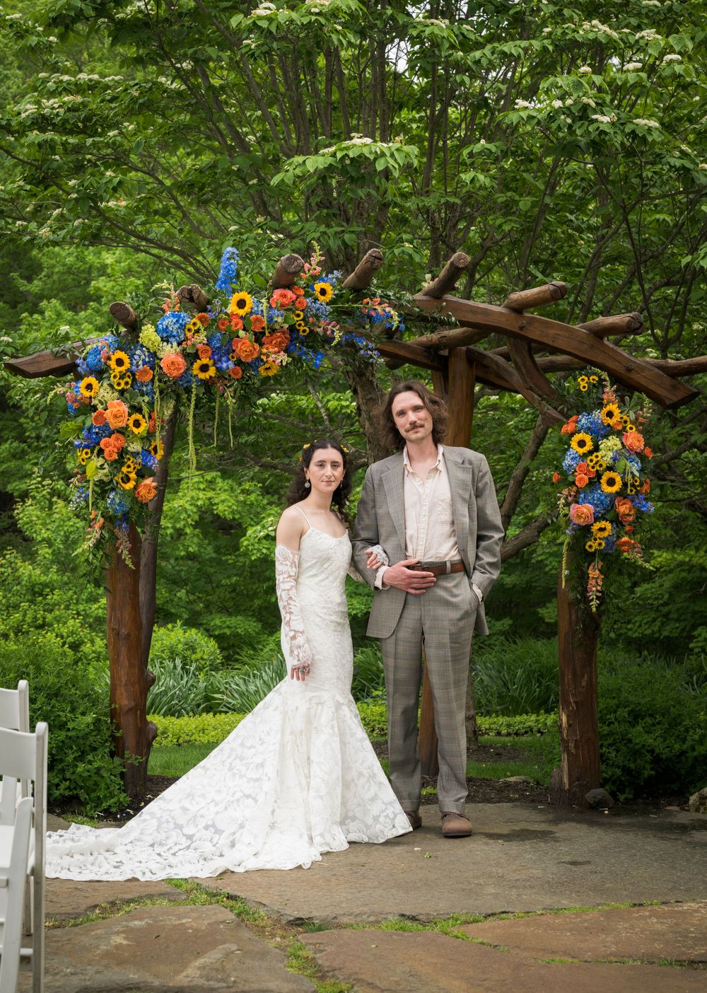Bride and groom pose under colorful floral arch with sunflowers in lush garden wedding ceremony.