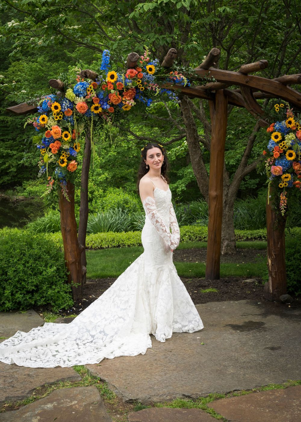 Bride in lace mermaid gown poses under rustic wooden arch adorned with sunflowers and colorful blooms in lush garden.