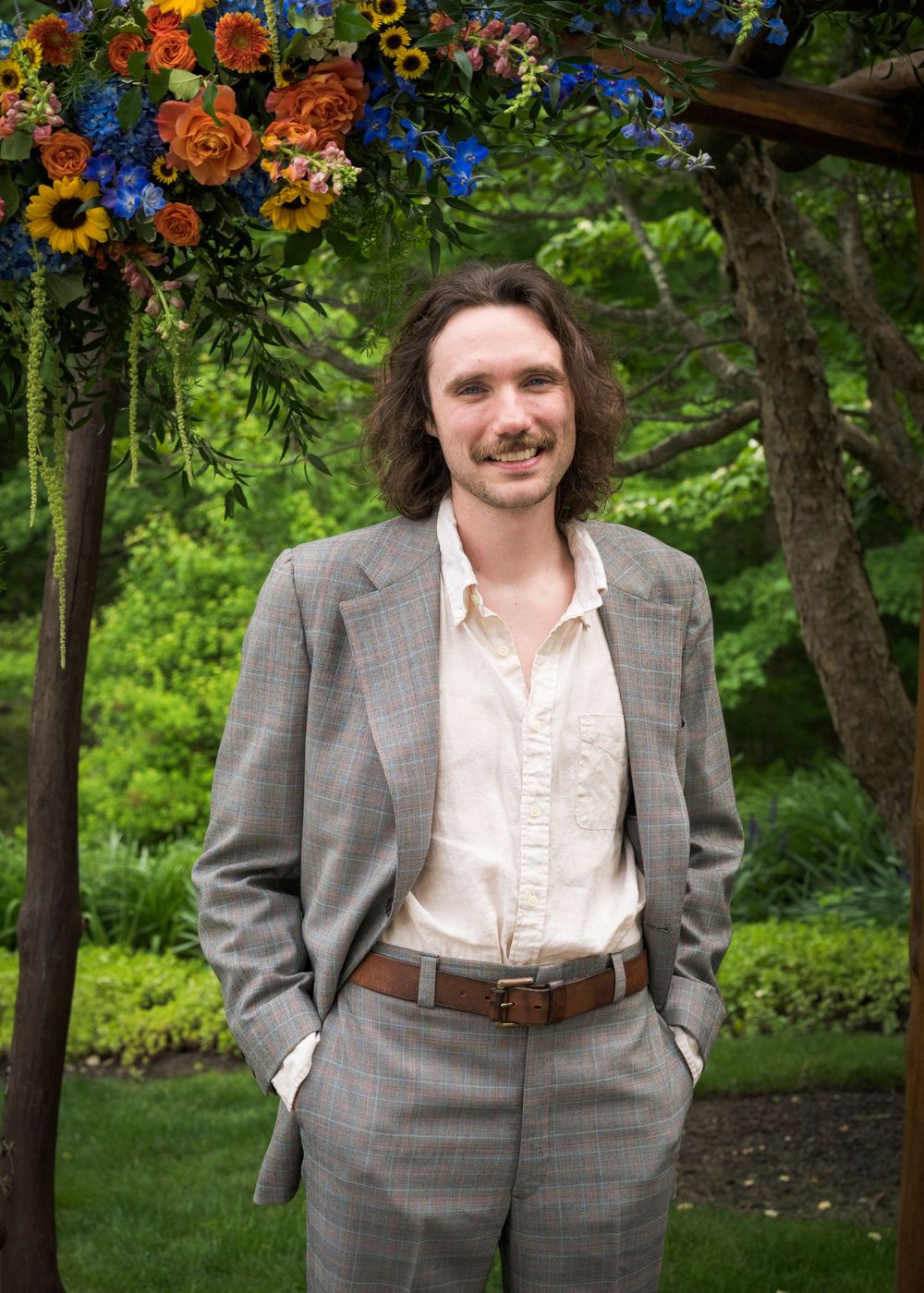 Young man with curly hair and mustache in gray suit smiling outdoors under colorful floral arch at wedding.