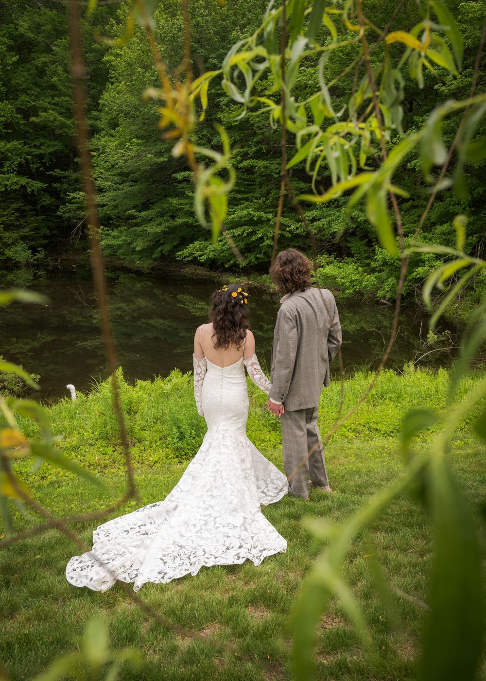 Bride and groom hold hands walking toward a serene river surrounded by lush green trees in outdoor wedding setting.