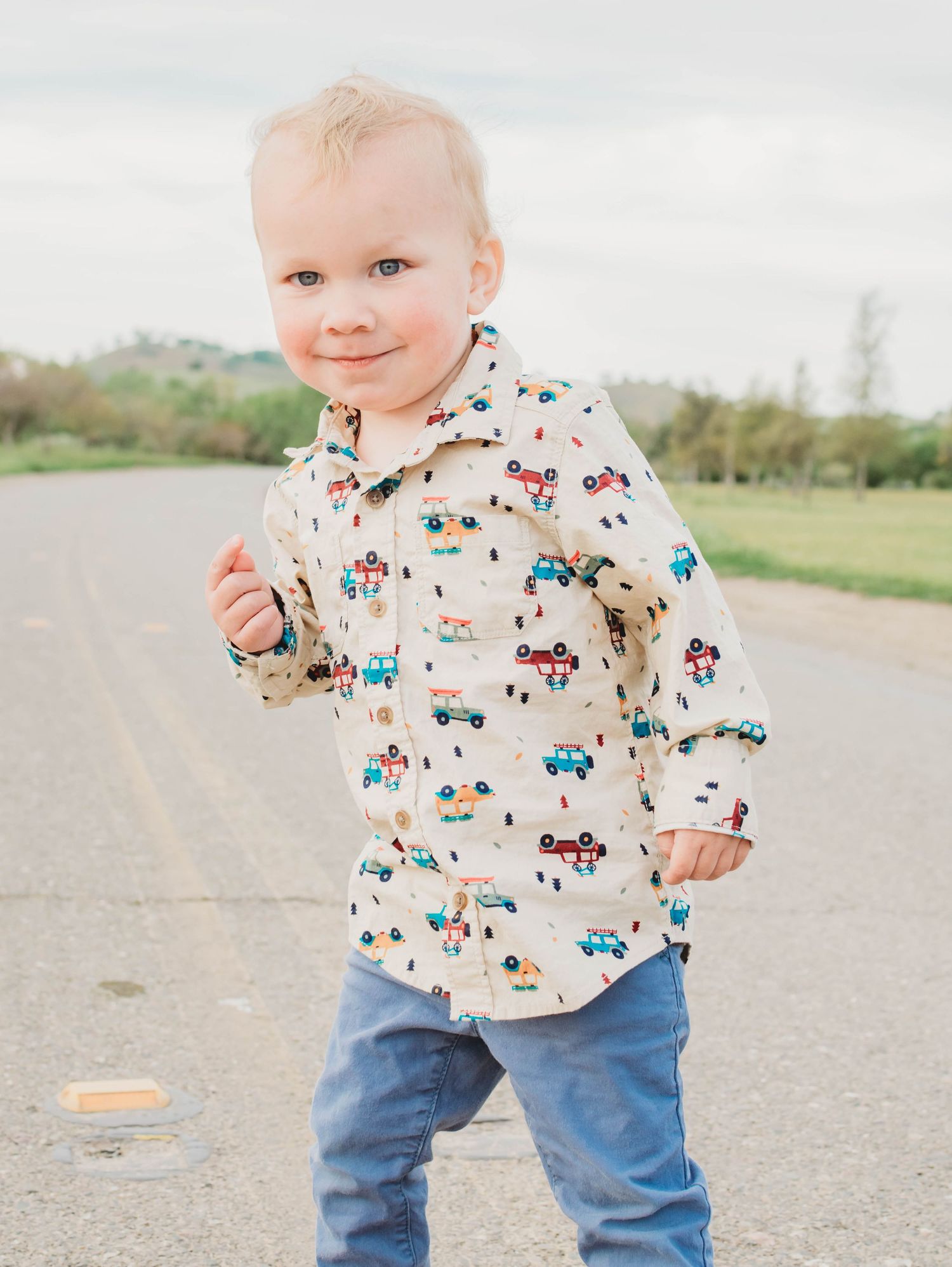 Toddler boy smiling in a printed button-up shirt and jeans, standing on a road outdoors.
