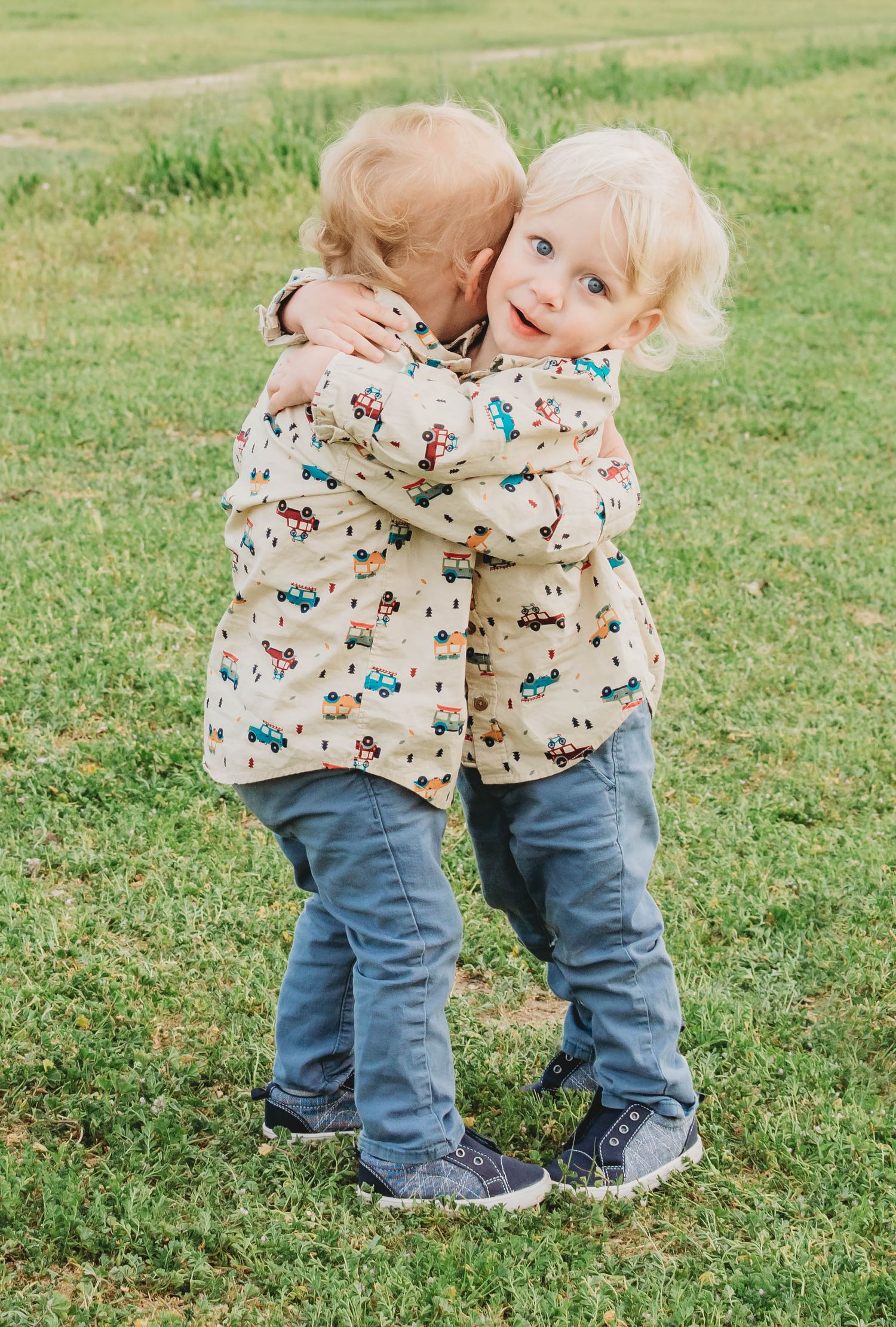 Two blonde toddlers in matching printed jackets and jeans hugging each other on a green grass lawn.