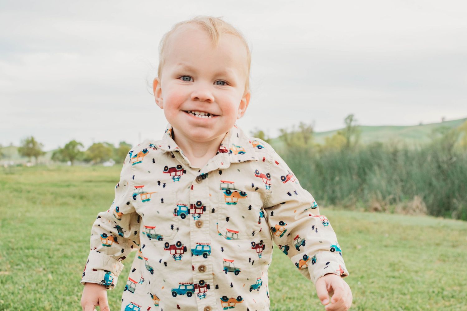 Smiling toddler in a printed shirt with vehicles runs outdoors on green grass under a cloudy sky.