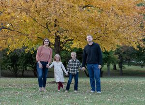 Wide landscape view of family members standing together in a park with golden fall foliage.