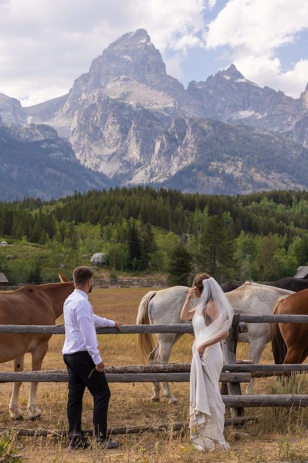 A couple stands near a wooden fence with horses against the dramatic backdrop of Grand Teton mountains in Wyoming.