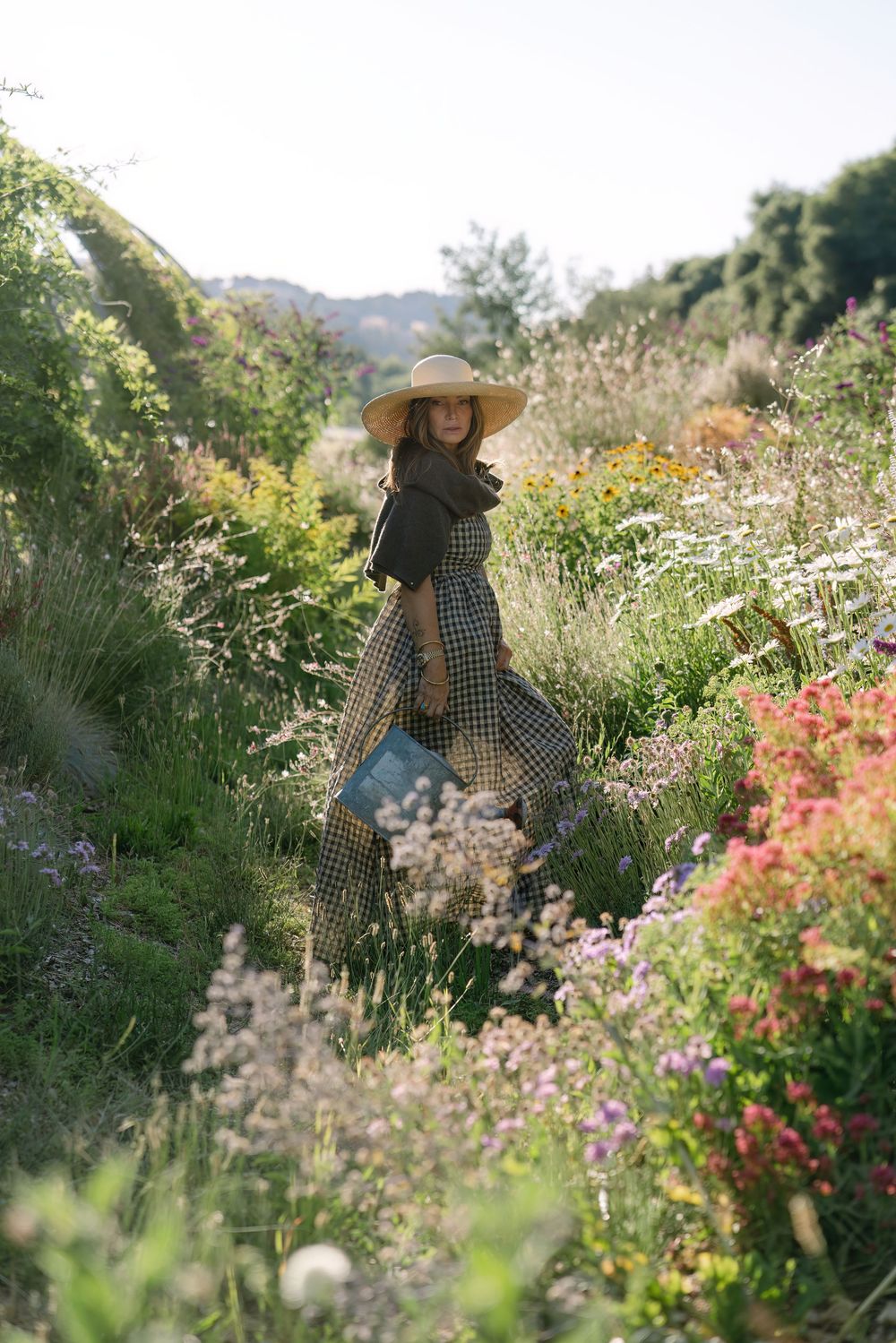 A sun-drenched garden scene with someone in a gingham dress amid blooming flowers.