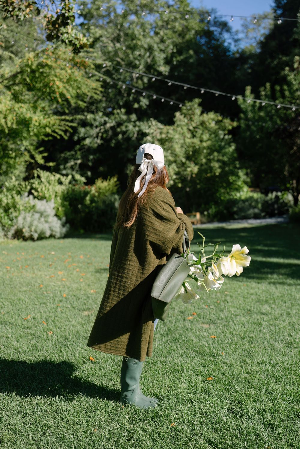 Person wearing olive coat and baseball cap walks across sunlit garden lawn.