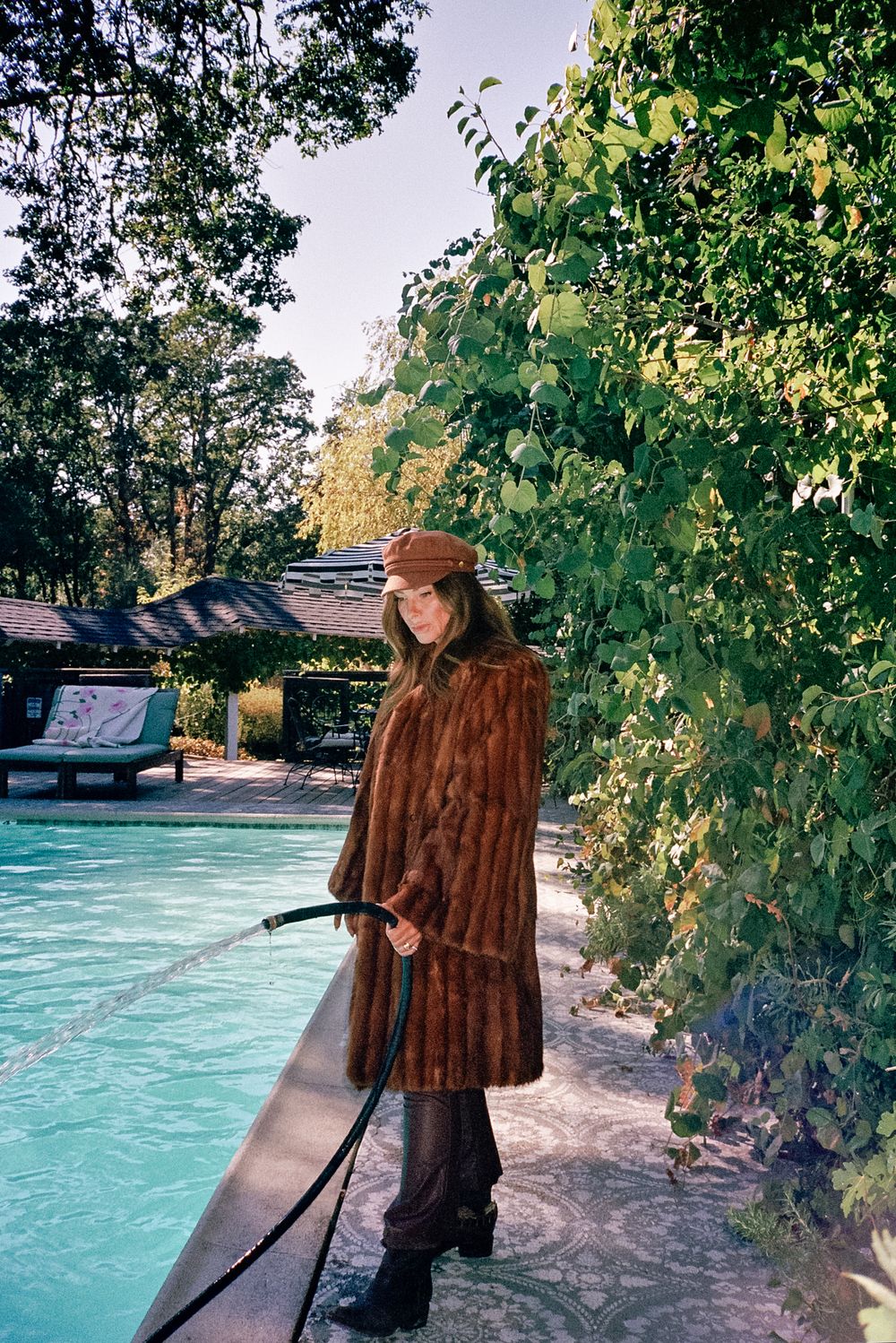 Person in brown coat and cap tends to swimming pool surrounded by green foliage.