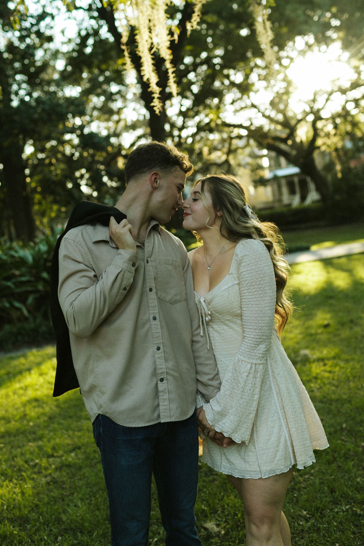 A man swings his jacket over one shoulder while leaning in to kiss his fiancee over the other
