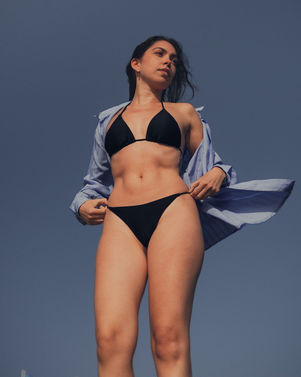 Person in black bikini and open light blue button-up shirt poses against blue sky in outdoor fashion photo.