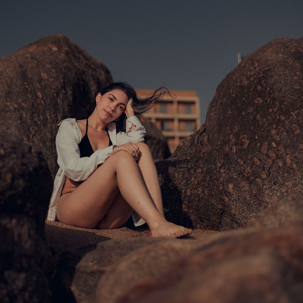 A person in beachwear poses thoughtfully on dark rocky terrain at dusk with moody lighting and dramatic shadows.
