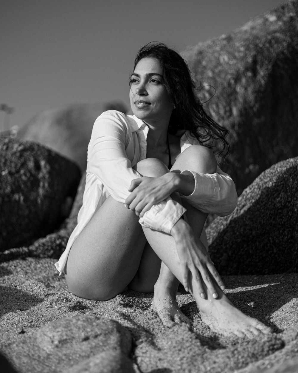 Black and white photo of someone in a white sweater sitting peacefully on coastal rocks in natural lighting.