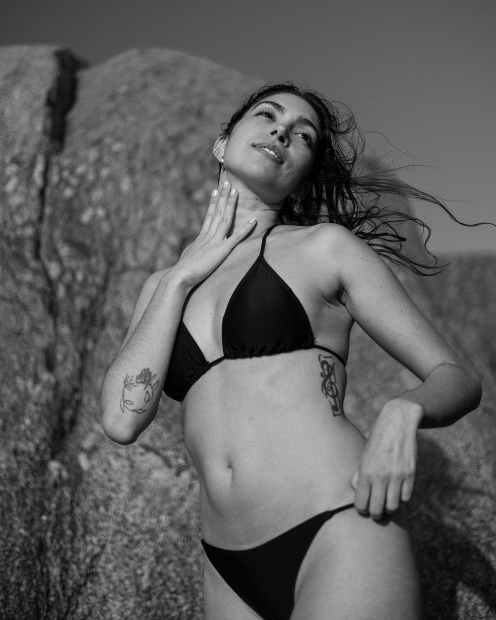 Black and white artistic portrait of someone in a black bikini posing dramatically against a rocky backdrop.