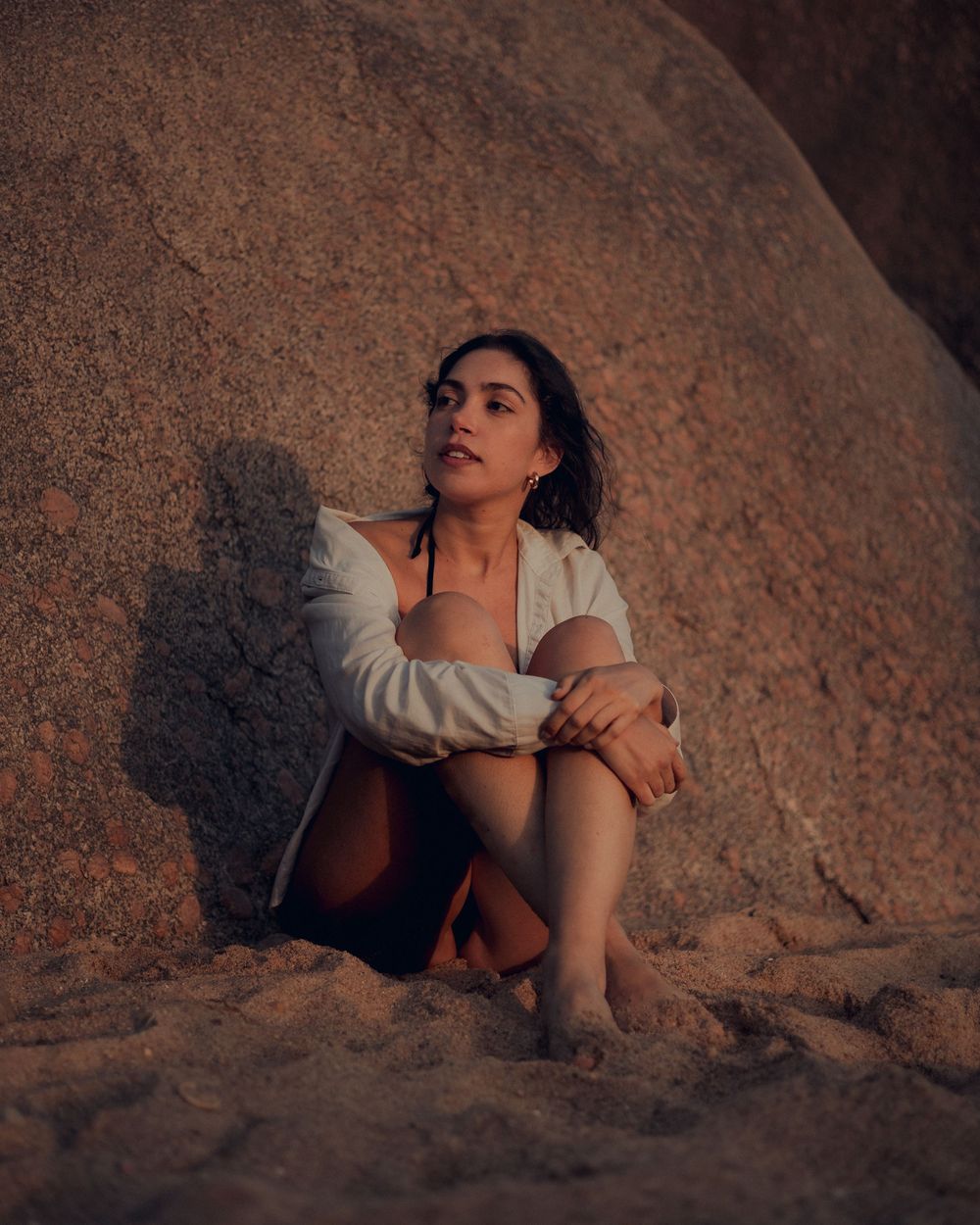A figure crouches in a contemplative pose against a textured rock formation at sunset creating a moody silhouette.