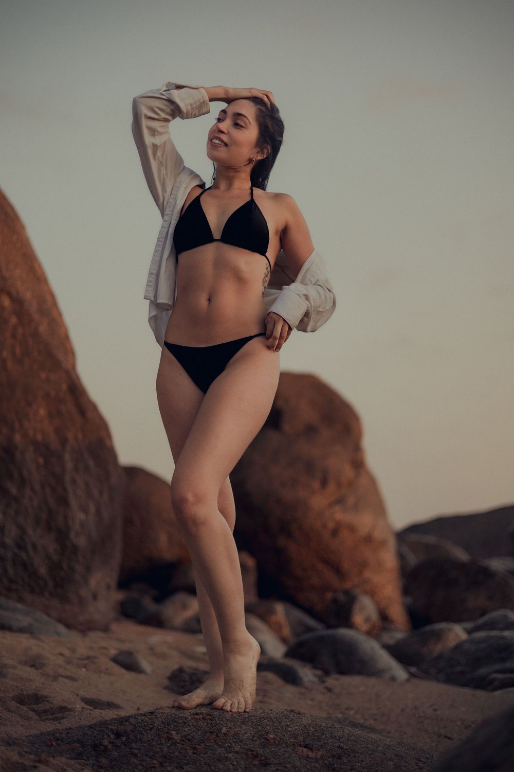 A person in a black bikini and light cardigan poses confidently on rocky coastal terrain at sunset.