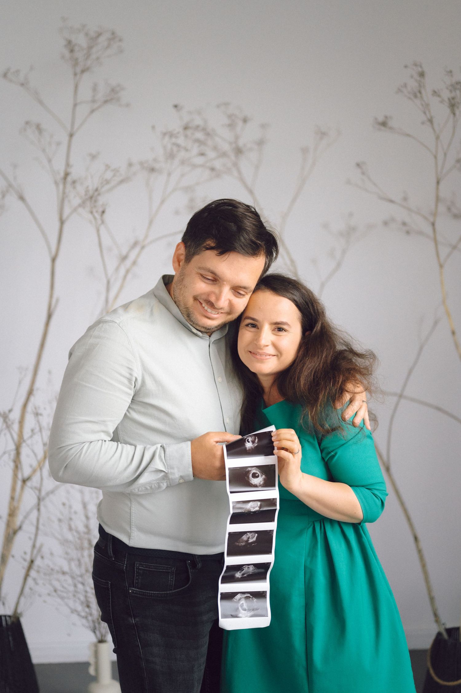 A couple in grey and green attire share a joyful moment holding ultrasound photos against a wall with floral decals.