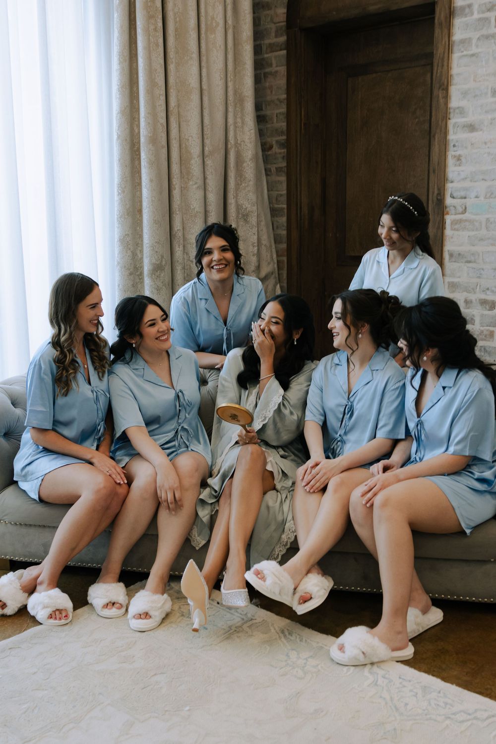 Bridesmaids in matching blue robes sit together on the floor sharing laughter and smiles during wedding preparations.