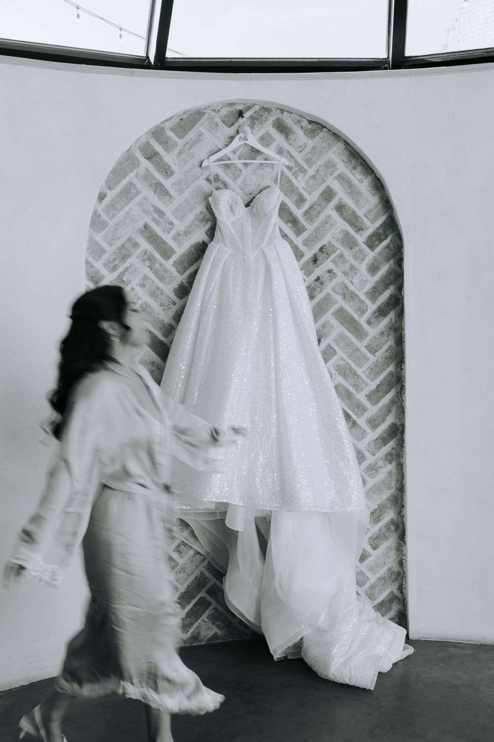 A flowing white wedding dress displayed in an elegant venue with a herringbone brick wall backdrop.