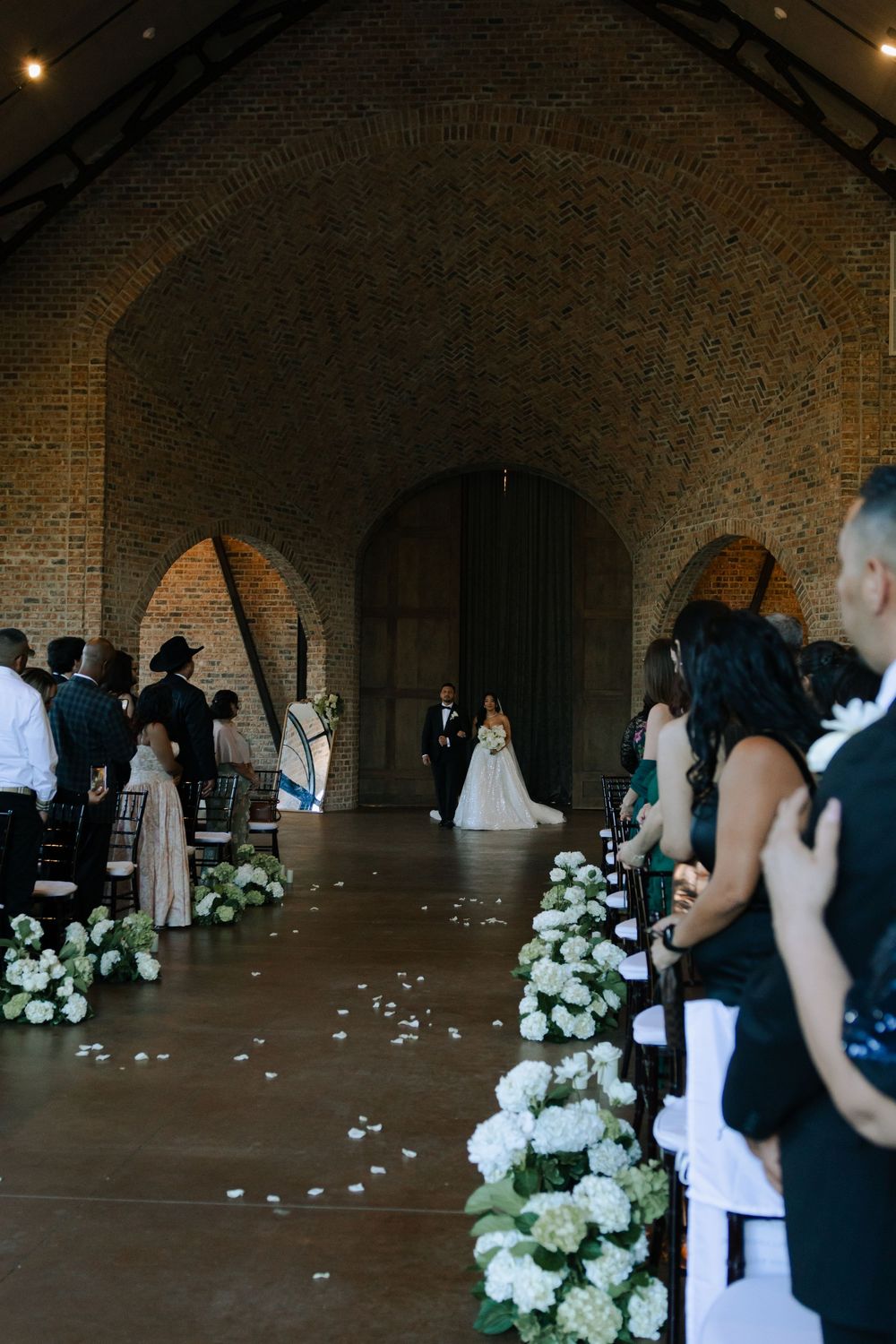 Wedding guests gather in a rustic brick-walled venue decorated with elegant white floral arrangements and scattered petals.