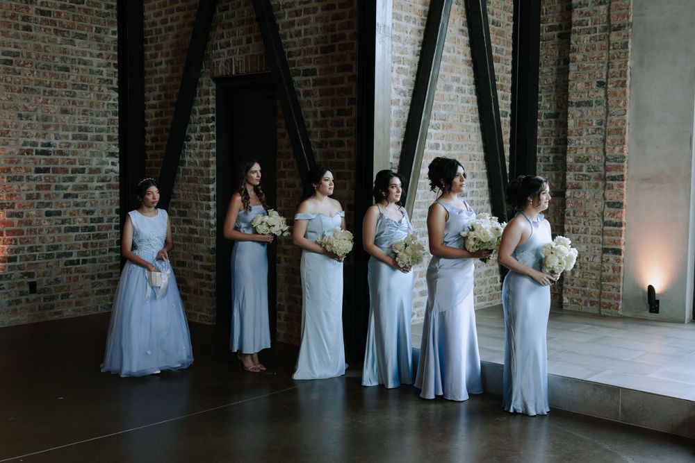 Wedding party in elegant white dresses and suits poses together against a rustic brick wall backdrop.