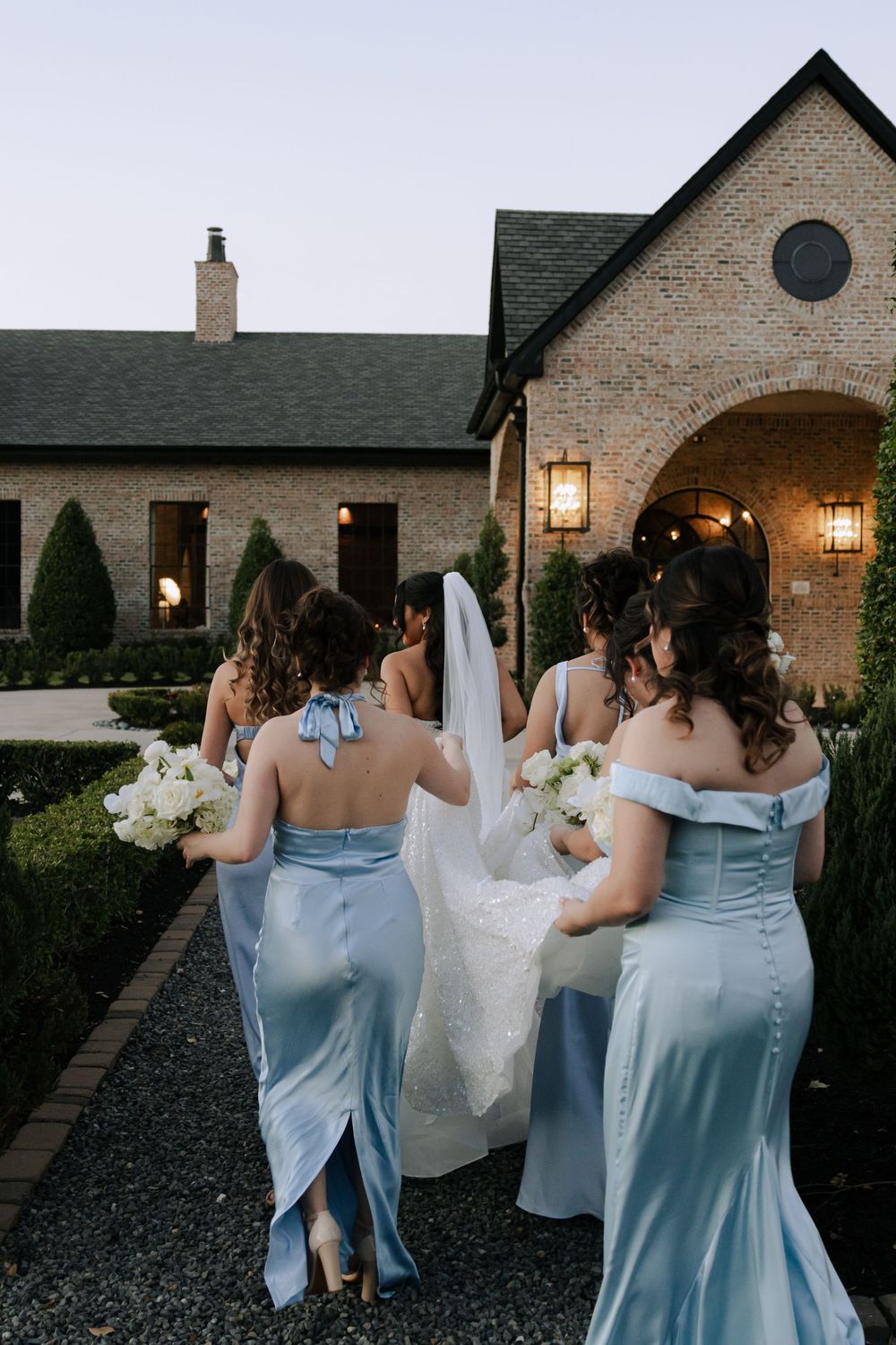Wedding party in light blue bridesmaid dresses walking together at dusk outside a brick venue with lanterns.