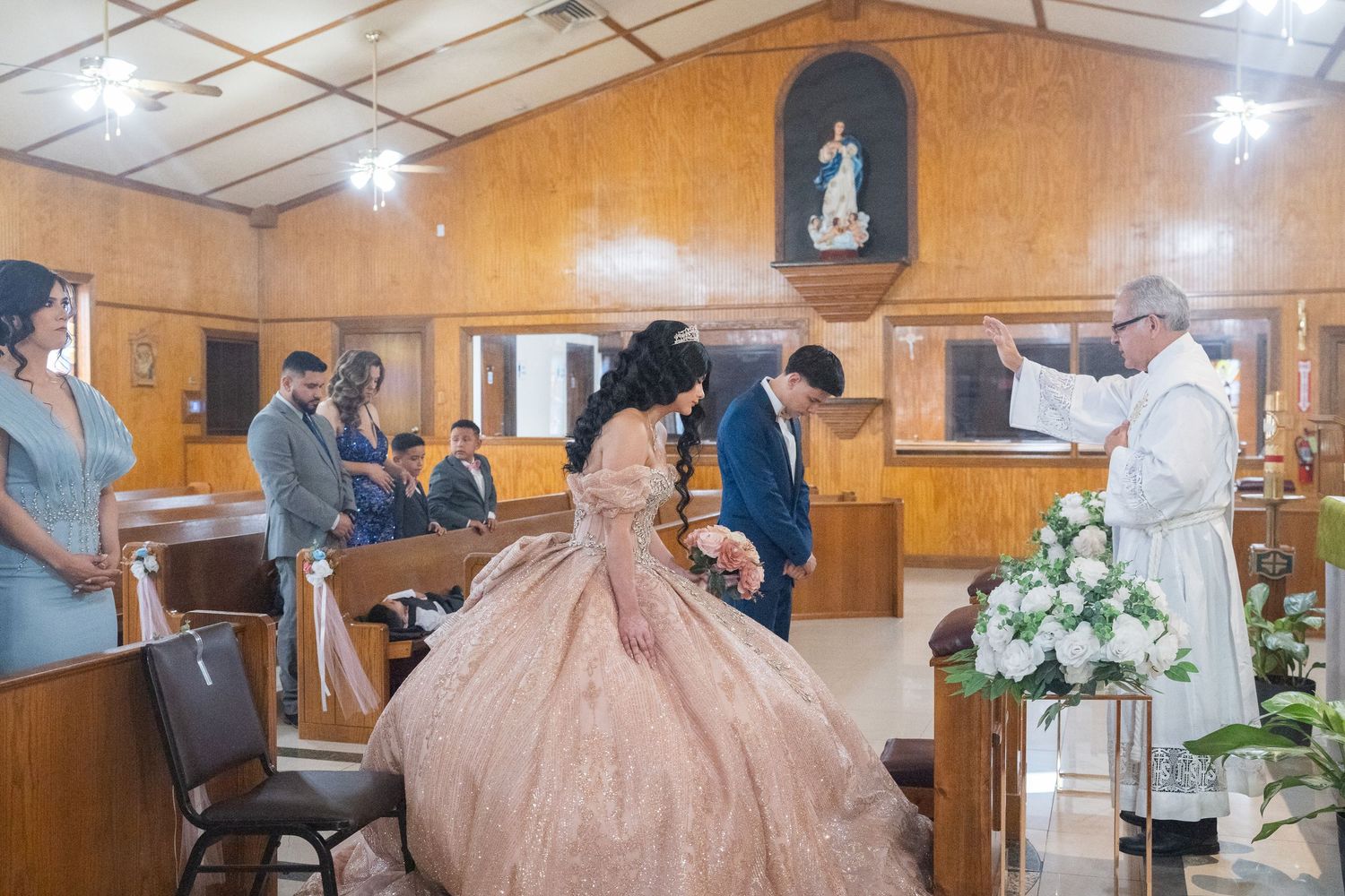 A quinceañera celebration in a wood-paneled church hall with floral decorations.