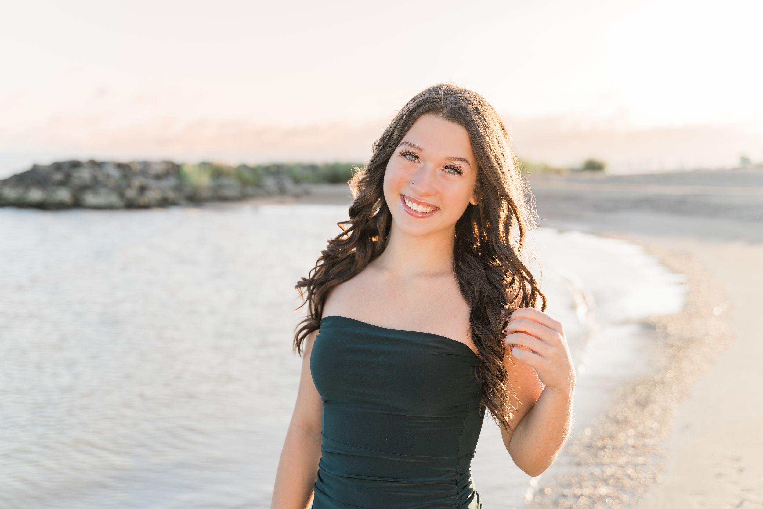 A woman in a black dress poses on a beach at sunset with golden light illuminating her figure.