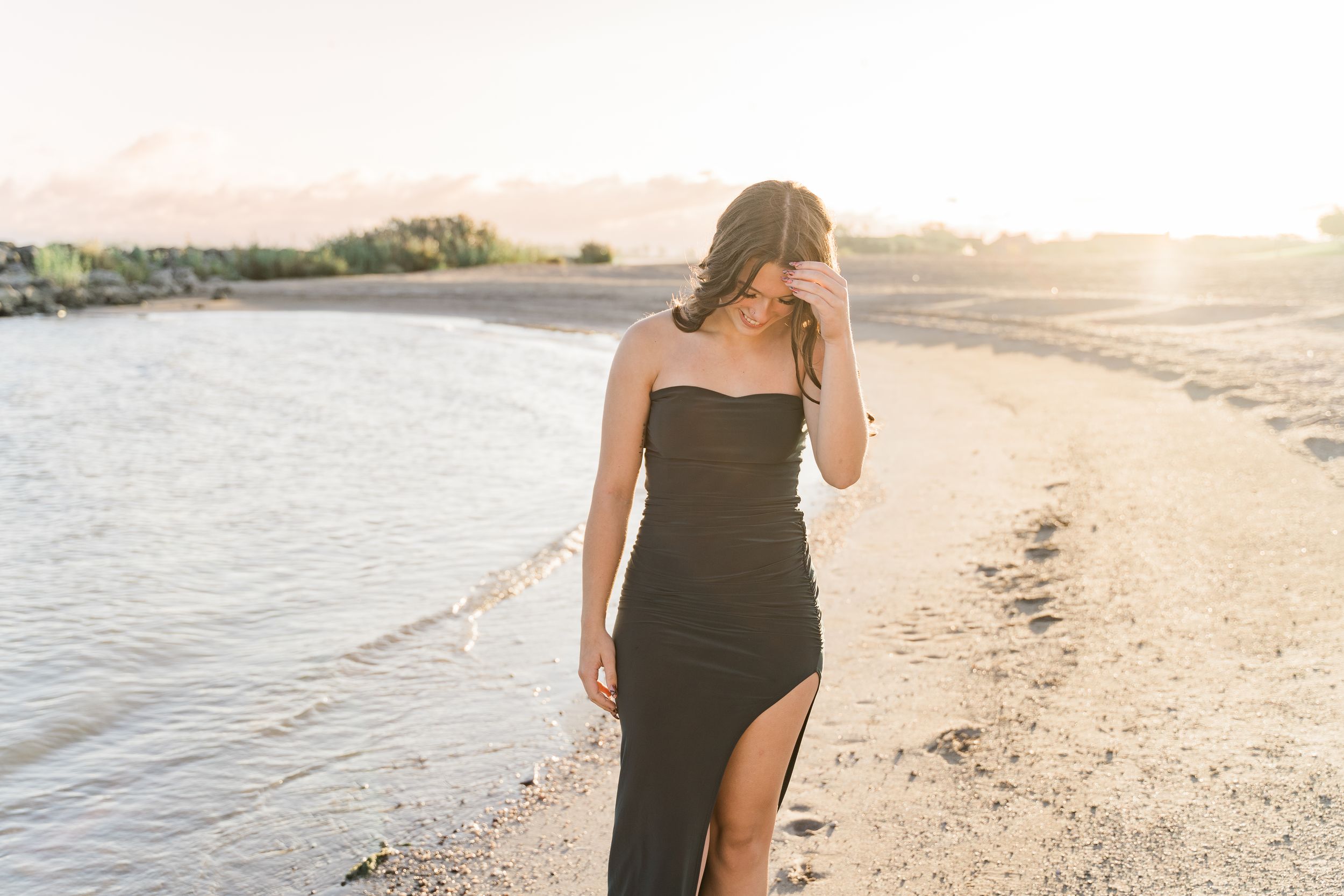 A woman in a black dress walks barefoot along a scenic beach shoreline at sunset.