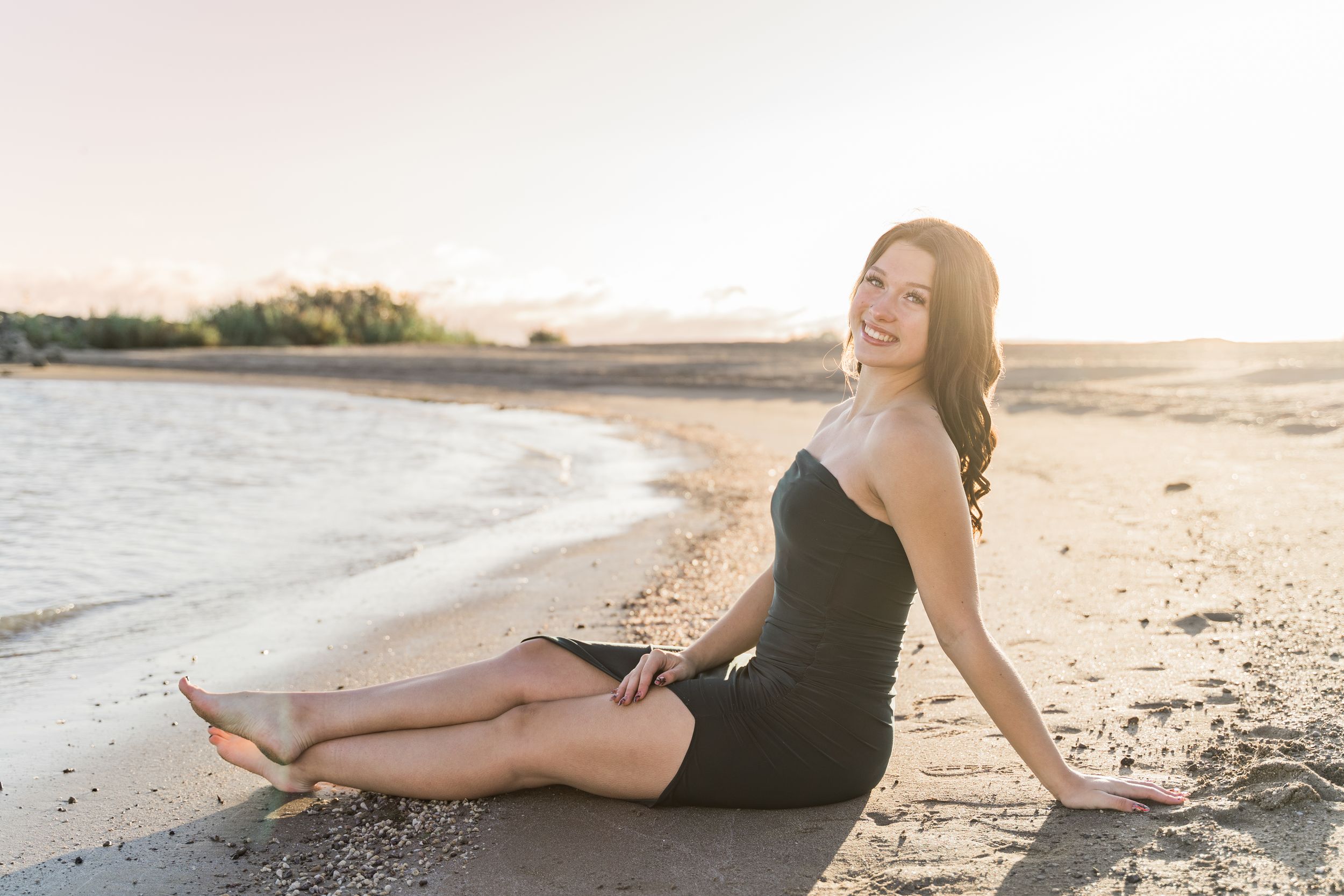 A woman in black dress lounges gracefully on a sun-drenched beach during golden hour with ocean waves in background.