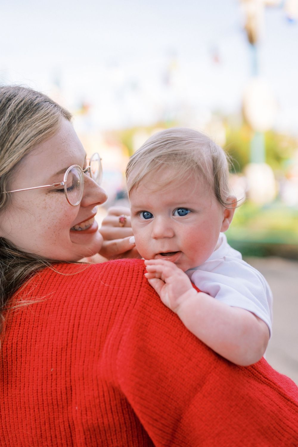 A tender moment between parent wearing a red sweater and baby with rosy cheeks against a soft bokeh background.