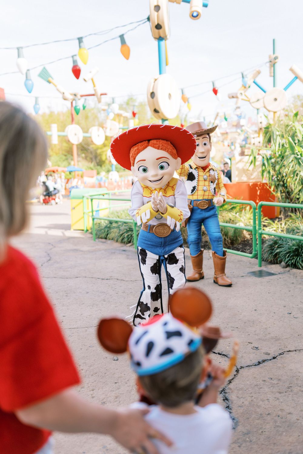 Toy Story characters Jessie and Woody stand in a theme park with colorful decorations and string lights overhead.