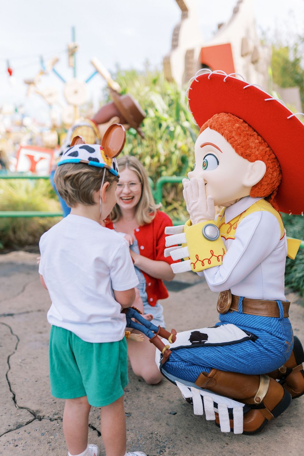 A toddler in green shorts interacts with a life-sized toy cowboy figure at a theme park.
