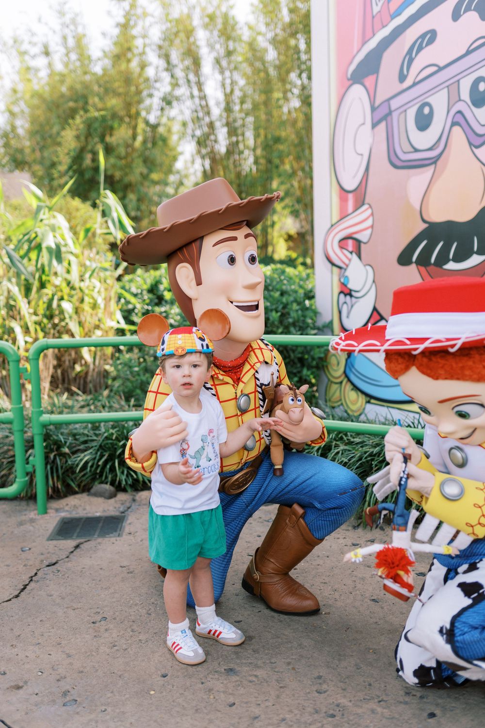 A costumed Woody character from Toy Story poses with a small child in green shorts at a Disney theme park.