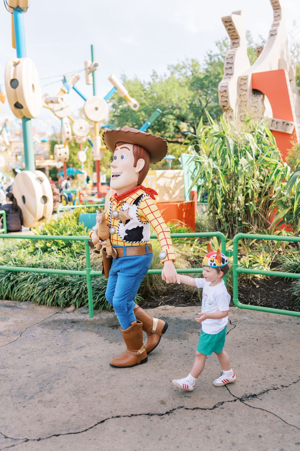 A Woody character from Toy Story interacts playfully with visitors in an outdoor park setting.