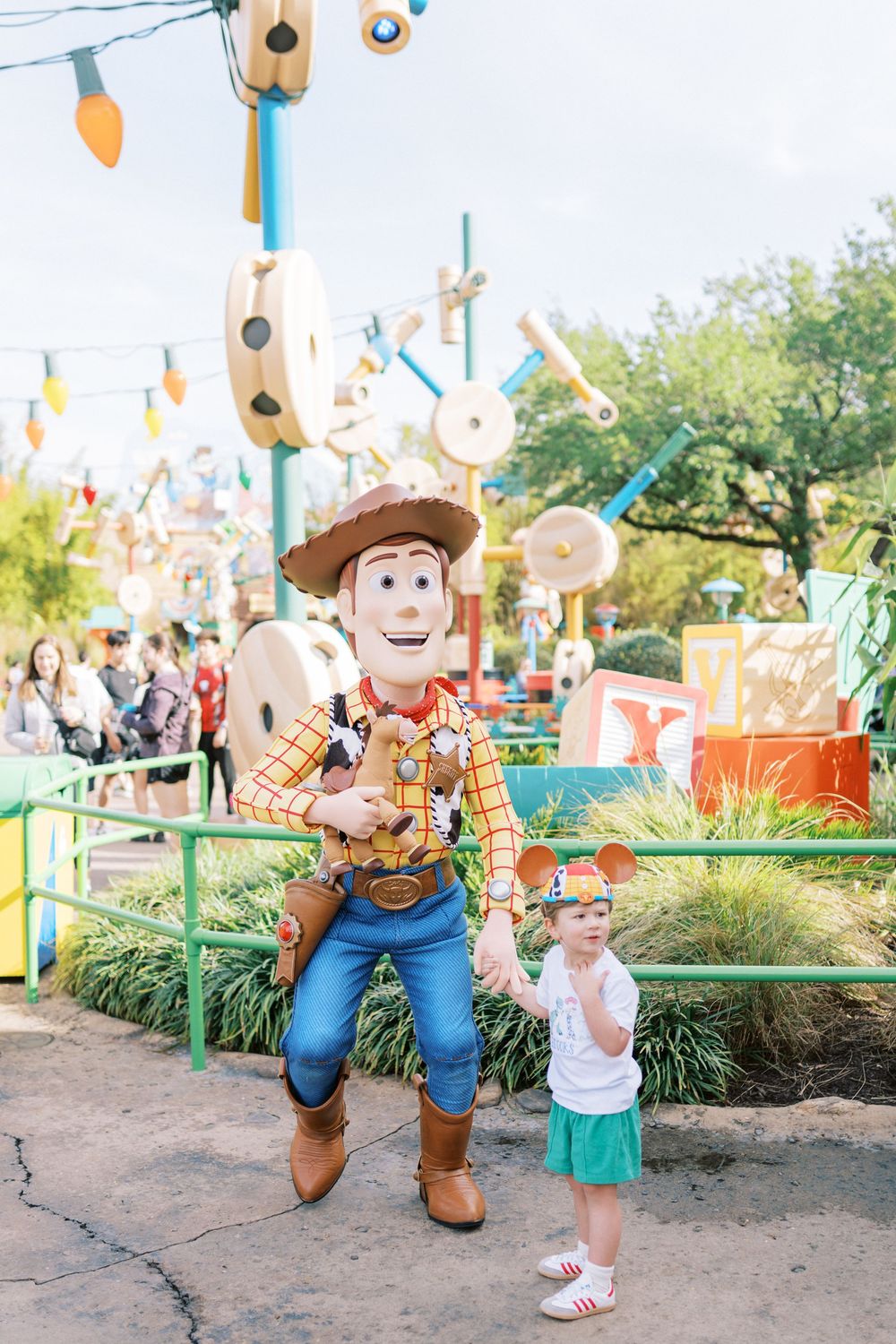 Theme park character dressed as a cowboy greets visitors under decorative string lights in a landscape setting.